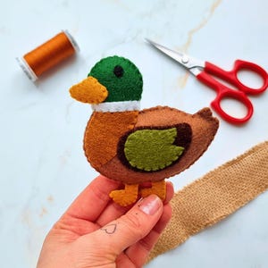May include: A handmade felt duck with a green head, brown body, and yellow beak and feet. The duck is held in a hand, with a spool of orange thread and red-handled scissors in the background. A piece of burlap ribbon is also visible.
