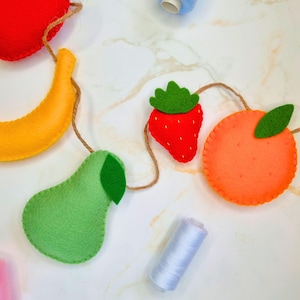 May include: A felt fruit garland with a red apple, yellow banana, green pear, red strawberry, and orange orange. The garland is strung with twine and has green felt leaves.