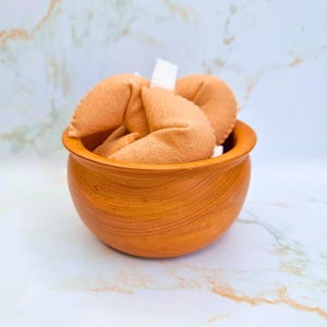 Puede incluir: Un cuenco de madera lleno de varias galletas de la fortuna de color marrón claro. El cuenco tiene un diseño liso y curvado y un color marrón cálido. Las galletas de la fortuna están hechas de un material suave y texturizado y tienen papel blanco asomando.