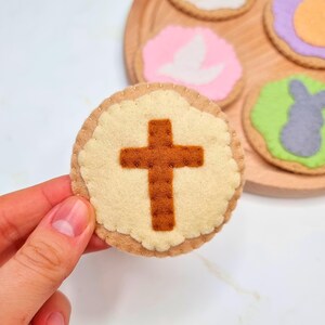May include: A felt Easter cookie with a brown cross on a cream background. The cookie is surrounded by other felt cookies with different designs.