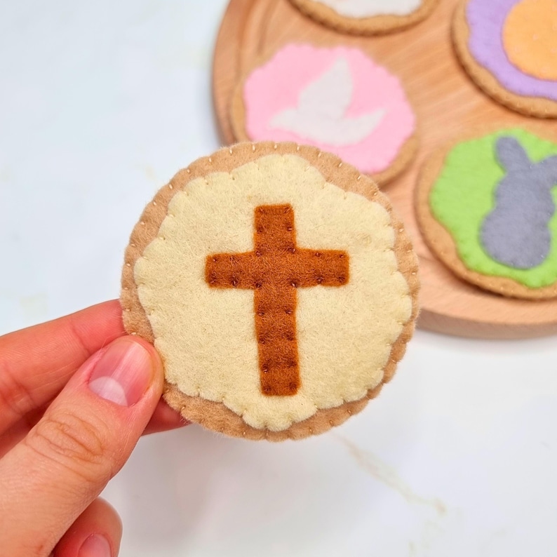 May include: A felt Easter cookie with a brown cross on a cream background. The cookie is surrounded by other felt cookies with different designs.