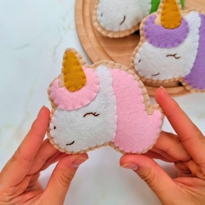 May include: A pink and white felt unicorn with a gold horn. The unicorn is being held by a person's hands.