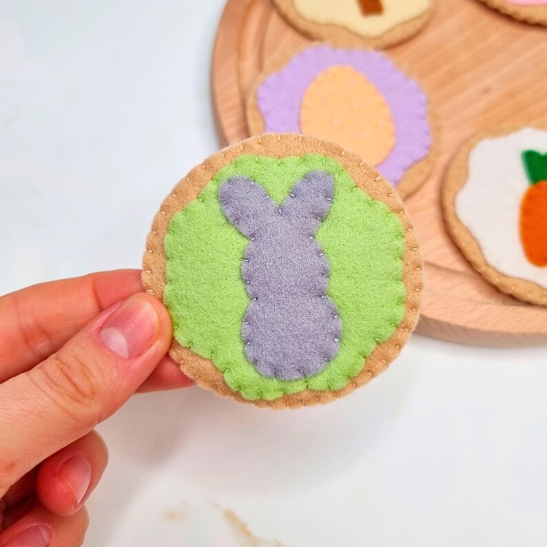May include: A felt cookie with a purple bunny on a green background. The cookie is surrounded by a brown felt border.