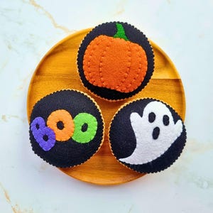 May include: Three circular felt ornaments featuring Halloween designs. One displays an orange pumpkin on a black background, another shows the word BOO in purple, orange, and green letters, and the third features a white ghost on a black background.