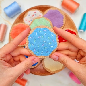 Puede incluir: Un conjunto de galletas de fieltro con diferentes glaseados y chispas de colores. Las galletas están dispuestas en un plato de madera.