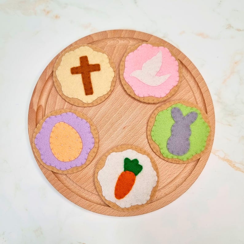 May include: Five felt Easter biscuits on a wooden board. The biscuits are decorated with a cross, a dove, an egg, a carrot and a bunny.