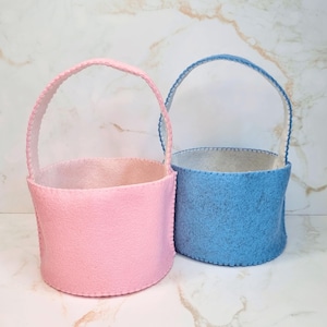 May include: Two round felt baskets with arched handles, one in a soft pastel pink and the other in a light blue. Each basket features visible stitched edges along the base and handle, creating a simple, structured cylindrical shape for storage.