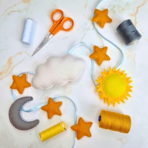 May include: A handmade celestial-themed mobile with felt stars, a sun, a moon, and a cloud, strung on a light blue ribbon. The image also shows orange scissors, spools of thread in white, gray, and yellow.