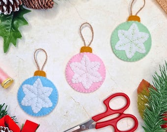 May include: Three circular felt ornaments featuring white stitched snowflake designs. One ornament is light blue, one is light pink, and one is light green. Each has a mustard yellow top cap and a brown twine loop for hanging, arranged with red scissors.
