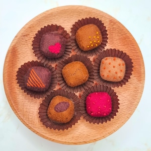 Peut inclure: Une assiette en bois présente sept chocolats en feutre dans des caissettes en papier marron. Les chocolats sont de différentes couleurs, dont le marron et le rouge, et décorés de détails brodés comme des cœurs, des points et des lignes.