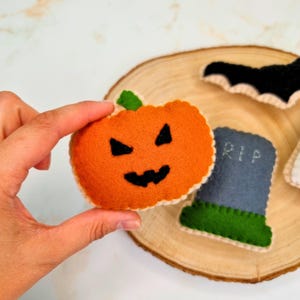May include: A felt Halloween decoration set featuring an orange pumpkin with a black stitched jack-o'-lantern face, a grey tombstone with the letters RIP stitched in white, and a black bat, all with visible blanket-stitched edges.