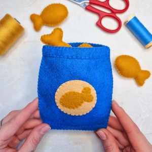 May include: A handmade blue felt bag with a beige fish-shaped emblem, filled with several yellow felt fish. Additional yellow felt fish are scattered around the bag. Sewing supplies, including scissors and thread spools, are visible.