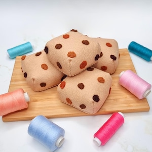 May include: Four felt brown and tan cookies with brown spots on a wooden cutting board surrounded by spools of colorful thread.