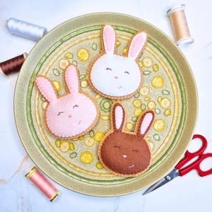 May include: Three felt bunny head ornaments in white, pink, and brown, each with pink inner ears and stitched facial features. They rest on a green and yellow patterned plate, with spools of thread and red scissors nearby. A craft-themed image.