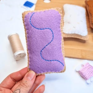 Może przedstawiać: Ręcznie robiony pop-tart z filcu z fioletowym nadzieniem i niebieskimi detalami. Prostokątny pop-tart ma jasnobrązową obwódkę. W tle widoczne są inne pop-tarty z filcu oraz szpulki nici.