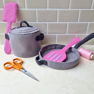 May include: A gray felt toy kitchen set with a pot, pan, and spatula. The pot has a lid and black handles. The pan has a black handle and a pink spatula. A pair of orange-handled scissors and a spool of pink thread are also present.