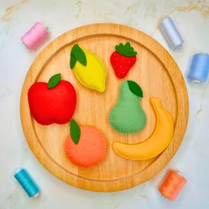 May include: A collection of felt fruit shapes including a red apple, yellow lemon, red strawberry, green pear, orange orange, and yellow banana arranged on a round wooden tray, surrounded by several small spools of colourful sewing thread.