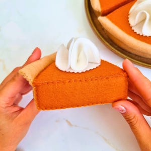 Op de afbeelding: Een vilten pompoentaartpunt met een topping van slagroom. De taart heeft een oranje vulling en een bruine korst. De punt wordt door twee handen vastgehouden, met een tweede taart op een bord op de achtergrond.