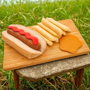 May include: A felt hot dog with red ketchup, fries, and a side of orange-colored food on a wooden cutting board. The board rests on a small stone table with wooden legs, set against a backdrop of green grass.