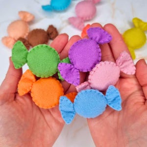 Op de afbeelding: Een verzameling kleurrijke vilten snoepjes in verschillende tinten, waaronder groen, paars, roze, oranje en blauw. Elk snoepje is rond met contrasterende, gewikkelde uiteinden. De snoepjes zijn handgemaakt en zien er zacht en pluche uit.