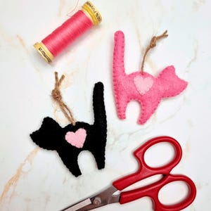 May include: Two felt cat ornaments, one black and one pink, each with a pink heart. A spool of pink thread and red scissors are also in the image. The ornaments have twine loops for hanging. The items are on a white marble surface.