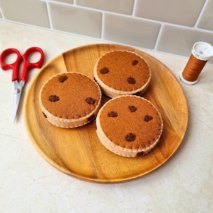 May include: Three felt chocolate chip cookies on a wooden plate. The cookies are brown with darker brown chocolate chips. A pair of red scissors and a spool of brown thread are next to the plate.