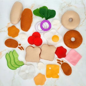 May include: A collection of felt play food items including two bread slices, a bagel, a donut, fried eggs, tomato slices, onion rings, green lettuce leaves, avocado slices, cheese slices, mushrooms, a meat patty, and a piece of ham.