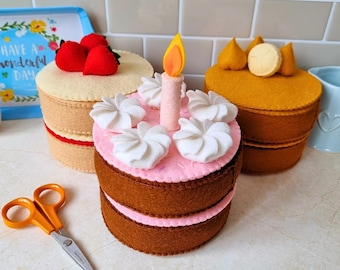 May include: Three round felt cakes with decorative toppings. One cake is beige with red strawberries, one is brown with pink frosting, white dollops, and a candle, and one is mustard yellow with yellow dollops and a round cookie topper.