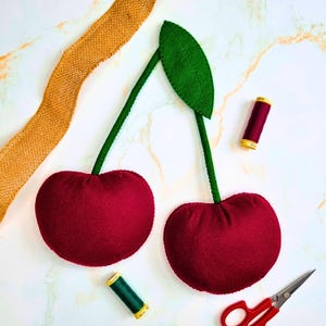 May include: Two plush cherry shapes made of dark red felt with green felt stems and a single green leaf. The cherries are accompanied by a spool of dark red thread, a spool of green thread, a pair of metal scissors, and a strip of gold burlap ribbon.