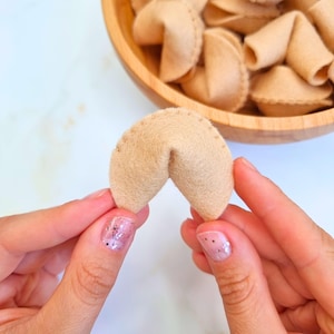 Puede incluir: Una galleta de la fortuna de fieltro beige se sostiene entre dos manos, con un cuenco de madera lleno de galletas similares en el fondo. La galleta tiene una costura cosida y una forma doblada. El fondo es de color claro y neutro.