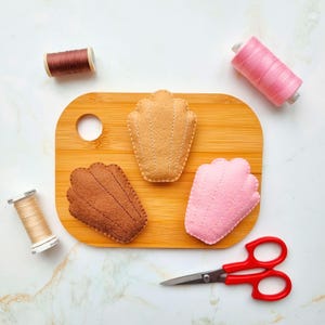 May include: A wooden chopping board with three felt seashell shapes in tan, brown, and pink. Spools of brown, pink, and beige thread are scattered around the board, along with a pair of red-handled scissors. The items are arranged on a white marble surface.
