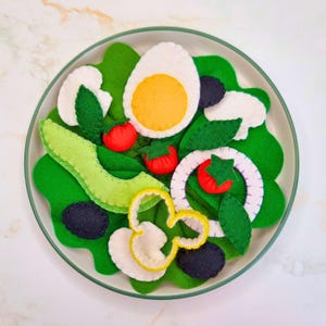 May include: A felt salad on a plate, with green lettuce, avocado slices, tomatoes, olives, a boiled egg, and onion rings. The plate has a green rim. The salad is a colourful, handcrafted item.