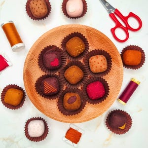 Peut inclure: Une assiette en bois présente des truffes en feutre dans des caissettes en papier marron. Les truffes sont de différentes couleurs, dont le marron, le rose et le rouge, avec des coutures décoratives. Des bobines de fil et des ciseaux rouges sont également visibles.