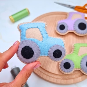 Könnte beinhalten: Handgefertigte Traktorspielzeuge aus Filz in Blau, Grün und Lila, mit grauen Rädern und schwarzen Zentren. Die Spielzeuge befinden sich auf einer Holzoberfläche, mit einer Spule grünen Fadens und einer Schere im Hintergrund. Die Traktorspielzeuge sind ein Bastelartikel.
