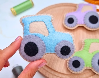 Può includere: Tre applicazioni in feltro a forma di trattore nei colori azzurro, lavanda e verde chiaro. Ogni trattore presenta un finestrino beige, ruote grigie con centri neri e visibili cuciture a festone beige lungo i bordi delle forme.