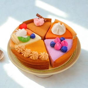 Könnte beinhalten: Ein Filz-Spielzeugkuchen, unterteilt in vier einzelne Stücke, mit verschiedenen Verzierungen wie einer roten Erdbeere, grüner Kiwi, blauen Blaubeeren, rosa Tupfen und weißen Sahnehäubchen. Der Kuchen ist auf einem runden, beigen Teller angerichtet.