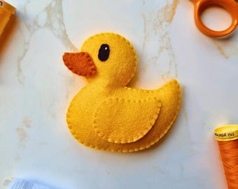 Puede incluir: Un pato de fieltro amarillo con un pico naranja texturizado y un ojo negro. El pato presenta bordes cosidos visibles a lo largo del cuerpo y el ala. Está rodeado por unas tijeras naranjas, un carrete de hilo naranja y un carrete de hilo blanco.