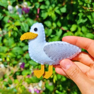 Op de afbeelding: Een handgemaakte vilten zeemeeuw met een witte body, grijze vleugels en gele snavel en poten. De vogel heeft een zwart oog en wordt vastgehouden tegen een achtergrond van groen gebladerte. Dit item is waarschijnlijk een decoratief ambacht.