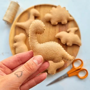 Peut inclure: Une découpe de dinosaure en feutrine beige avec des bords cousus visibles tenue au premier plan. Plusieurs autres formes de dinosaures en feutrine beige sont disposées sur un plateau rond en bois, avec une bobine de fil beige et des ciseaux en métal à poignées orange.