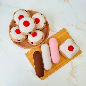 Puede incluir: Un conjunto de alimentos de fieltro, que incluye seis galletas redondas con glaseado rojo y blanco y cuatro pasteles largos y rectangulares. Las galletas están dispuestas en un plato de madera, mientras que los pasteles están en una tabla de cortar de madera.