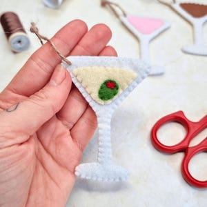 May include: A felt martini glass ornament featuring a white frame, a pale yellow liquid center, and a green olive with a red pimento center. The ornament includes a brown twine loop for hanging and is constructed with visible white stitching.