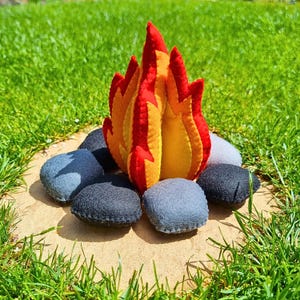 May include: A plush felt campfire toy featuring layered flame shapes in vibrant red, orange, and yellow. The flames are surrounded by a circular arrangement of soft, stuffed felt stones in shades of dark charcoal gray and light heather gray.