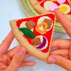Puede incluir: Una porción de pizza de fieltro con salsa roja, corteza beige y varios ingredientes. Los ingredientes incluyen trozos de fieltro marrón, naranja, verde y morado. La porción de pizza está sostenida por dos manos, con una pizza más grande en el fondo.