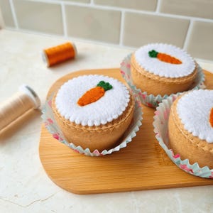 May include: Three felt carrot cake cupcakes on a wooden board. Each cupcake has a white felt top with a felt carrot and green leaf detail. The cupcakes sit in decorative paper liners. A spool of orange thread and a spool of white thread are in the background.