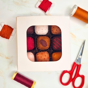 May include: A box of nine felt chocolates in a variety of colors and textures, displayed in a white box with a square cutout. Red scissors, spools of red and orange thread, and a spool of burgundy thread are arranged around the box.