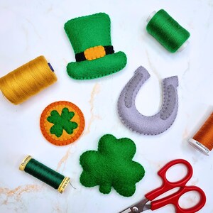 May include: A collection of St. Patrick's Day themed felt decorations, including a green leprechaun hat, a horseshoe, a four-leaf clover, and a gold coin with a shamrock. Spools of green, gold, and orange thread and red scissors are also present.