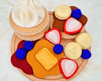May include: A felt play food set featuring a stack of pancakes topped with white cream, a pancake with brown spread, banana slices, strawberries, and blueberries, plus a separate pancake with a yellow butter pat and fruit pieces on a wooden tray.