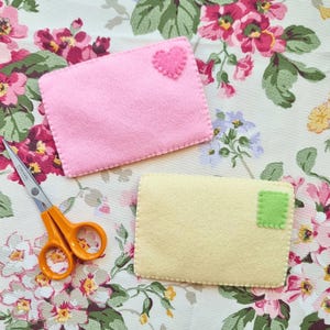 May include: A light pink felt envelope with a small pink heart appliqué and a pale yellow felt envelope with a small green square appliqué, accompanied by a pair of orange-handled metal scissors with silver blades and a visible screw.