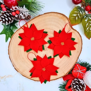 Könnte beinhalten: Drei rote Weihnachtssterne aus Filz mit grünen Filzblättern und kleinen goldenen Perlen in der Mitte. Jede Blume hat eine dünne rote Aufhängeschlaufe an der Oberseite und ist auf einer runden Holzscheibe mit natürlicher Rinde angeordnet.