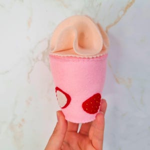 Puede incluir: Un batido de fresa de fieltro rosa con una cobertura de crema batida de fieltro rosa. El batido tiene dos rodajas de fresa en el lateral.